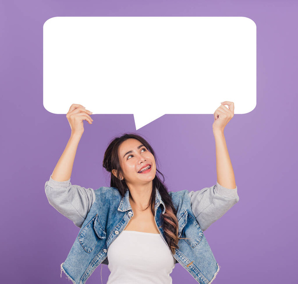 Happy Asian beautiful young woman smiling excited wear denims hold empty speech bubble sign, Portrait female posing show up for your idea looking at bubble, studio shot isolated on purple background - Foto, Bild