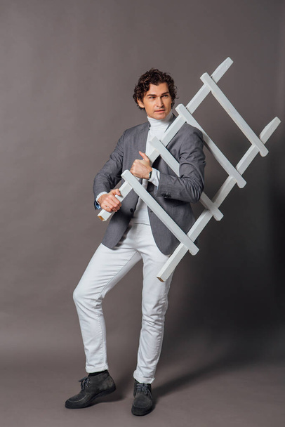 Fashion portrait of a tall handsome man dressed in white turtleneck, white jeans and grey jacket posing on the grey background with white ladder - Fotó, kép