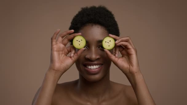Mujer africana posando con rodajas de pepino cubriendo los ojos, fondo beige - Imágenes, Vídeo