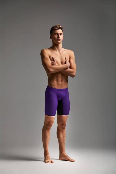 Młoda atrakcyjna pływaczka w doskonałej formie fizycznej w fioletowych spodenkach kąpielowych, na szarym tle, przestrzeń do kopiowania - Zdjęcie, obraz