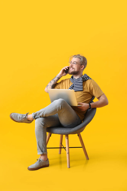 Young man with laptop and phone sitting in armchair on color background - Foto, Bild