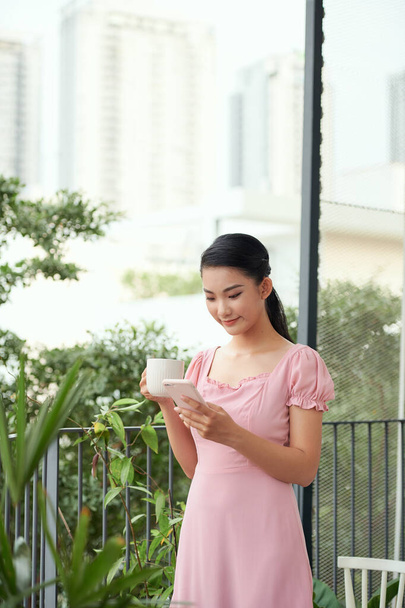 Ευτυχισμένη Ασιάτισσα στο μπαλκόνι κρατώντας την κούπα του καφέ, διαβάζοντας μήνυμα από το κινητό της τηλέφωνο και χαμογελώντας - Φωτογραφία, εικόνα