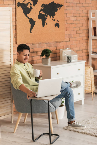 Fiatal afro-amerikai srác laptoppal kávét iszik, miközben otthon ül karosszékben. - Fotó, kép