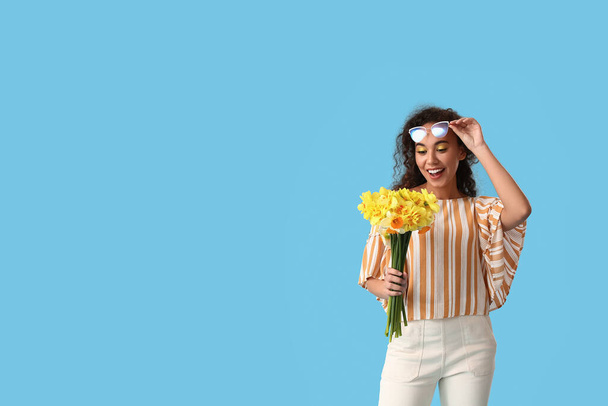 Surprised young African-American woman with narcissus bouquet on color background - 写真・画像
