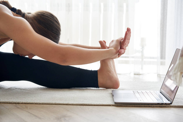 Mujer joven practicando yoga, sentada en ejercicio de flexión hacia adelante sentada, pose paschimottanasana, ejercitándose, usando ropa deportiva - Foto, Imagen