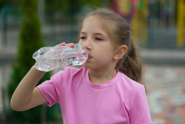 Egy szőke, hosszú hajú lány rózsaszín pólóban vizet iszik egy üvegből a parkban lévő játszótéren. Egészséges életmód koncepció. Szelektív fókusz - Fotó, kép