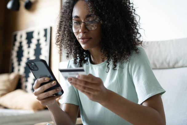 Happy millennial mixed race woman holding banking credit card, entering payment information in smartphone application - Zdjęcie, obraz