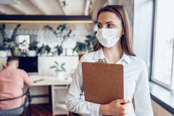 Young businesswoman in face mask standing in office and holding clipboard with documents - Photo, image