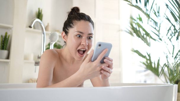 Excited young multi ethnic asian woman celebrating bid win success victory or getting ecommerce shopping offer on smartphone, during morning daily body care and beauty procedure in bathroom - 写真・画像