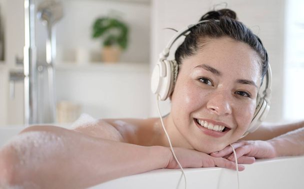 Attractive smiling young multicultural asian woman with headphones enjoying a hot bath. Multi ethnic millennial girl listening music relaxing in bathroom, treatment therapy healthy lifestyle concept - Foto, Imagem