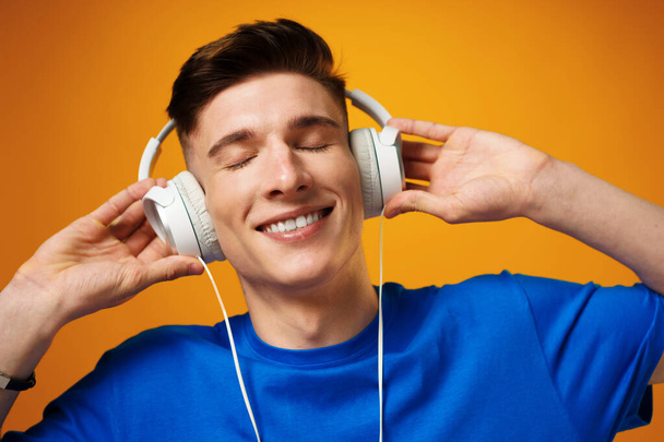 Young man in blue t-shirt listening to music with headphones against yellow background - Фото, изображение