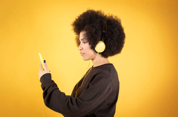 mooie afro vrouw luisteren naar muziek met koptelefoon en smartphone, gele achtergrond - Foto, afbeelding