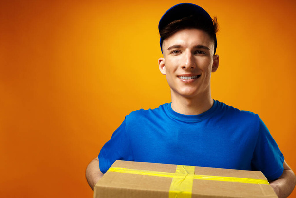 Young handsome delivery man holding parcel cardboard box against yellow background - Foto, Imagen