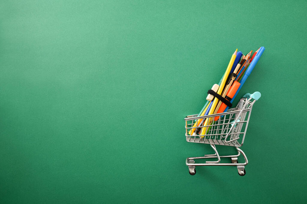Purchase of school tools with cart full of supplies on green background. Top view. - Foto, afbeelding
