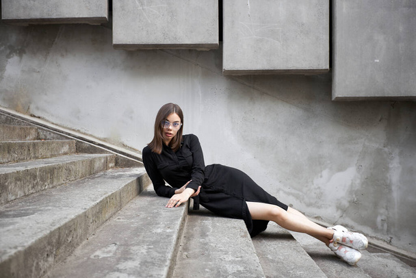 Fashion portrait of a sexy brunette woman with a make up wearing black clothes. She is wearing black modern dress, white sneakers and glasses - Fotografie, Obrázek