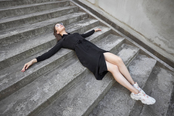 Szexi barna nő egy smink visel fekete modern divatos ruha feküdt cement lépcsőn - Fotó, kép