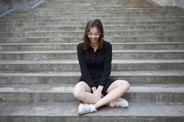 Woman in black dress sitting on cement stairs in summer in urban city area. She is happy and relaxed - 写真・画像
