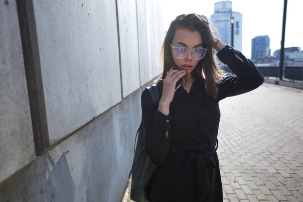 Business woman portrait in city streets at sunset. Sexy brunette wearing black dress, glasses and black leather bag outdoor - Φωτογραφία, εικόνα
