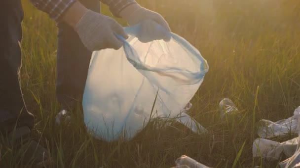 Un hombre con guantes protectores recoge basura con las manos en una bolsa de basura, trabajo voluntario en la naturaleza al aire libre, limpieza de botellas de plástico, vasos, servilletas en hierba verde, eco, contaminación de la naturaleza - Metraje, vídeo