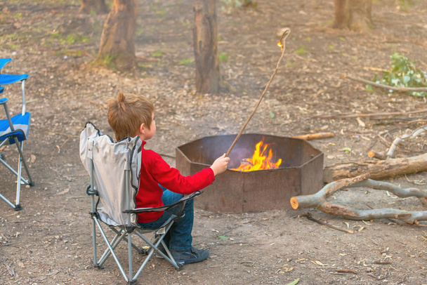 Boy burning a stick in the camp fire while sitting in the chair during winter school holidays, Kuitpo forest, South Australia - Photo, Image