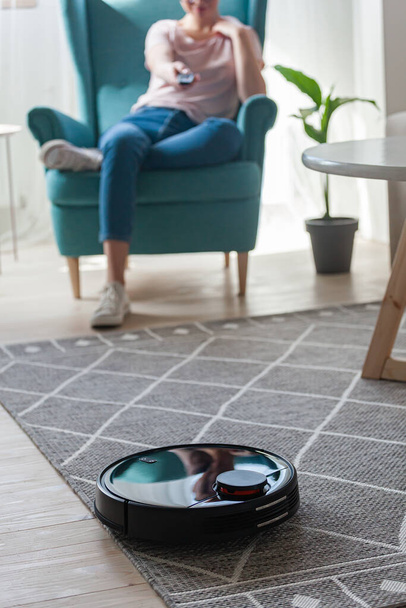Robotic vacuum cleaner cleaning carpet, woman controls remote control and joy rest while sitting on armchair at home - 写真・画像