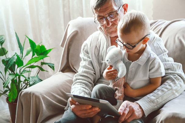 An elderly grandfather and his little grandson are using a tablet device together, smiling and having fun or watching a funny video on the Internet, sitting in a chair - Photo, Image