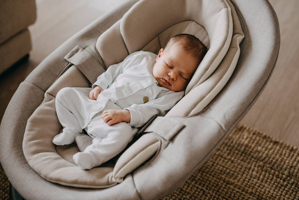 Newborn baby sleeping in a swing, wearing a white onesie pajama. - 写真・画像