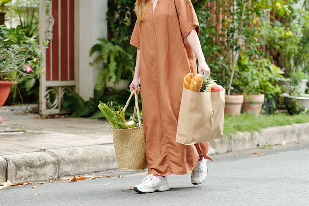Zugeschnittenes Bild einer jungen Frau, die zwei Taschen voller Lebensmittel, Brot, Gemüse und Obst trägt - Foto, Bild