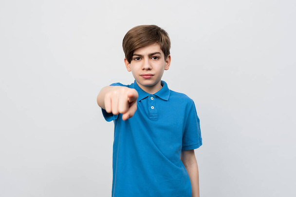 Серьезный мальчик 12-14 лет в повседневной синей футболке, указывая пальцем на камеру и на вас, уверенный в себе жест выглядит серьезным. Студийный кадр, серый фон, изолированный - Фото, изображение