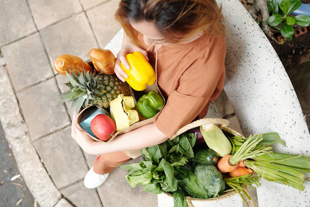 Mooie jonge vrouw zittend op de bank met twee grote zakken verse groenten, fruit en brood - Foto, afbeelding