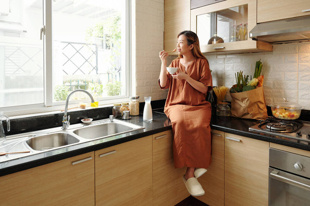 Positiivinen kaunis nuori nainen istuu keittiön tiskillä, syö maissihiutaleita ja katselee ikkunasta - Valokuva, kuva