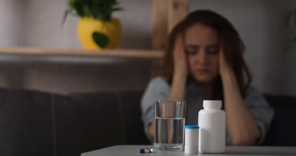 Nuori nainen koskettaa migreenistä kärsiviä ohimojaan. Joitakin pillereitä edessä pöydällä. - Materiaali, video