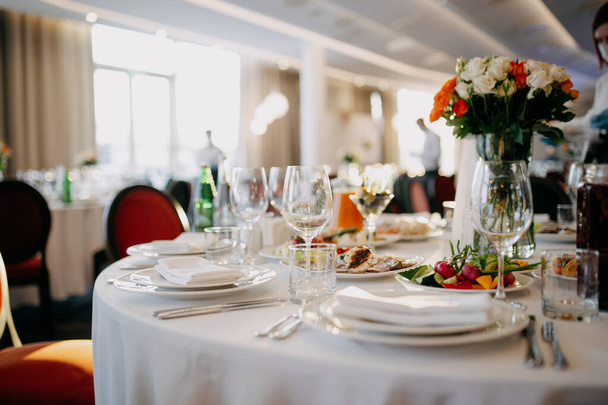 Готовлю свадебный стол. Ресторанный бизнес. Очки, белые тарелки и столовые приборы на круглом столе, украшенном розами в белой скатерти. - Фото, изображение