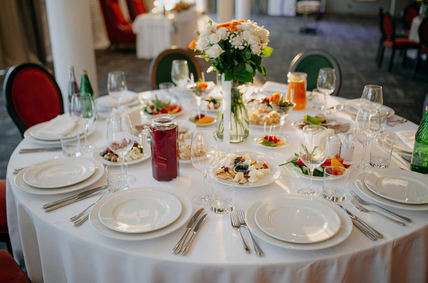 Tarjoilen hääpöytää. Ravintola-alalla. Lasit, valkoiset lautaset ja ruokailuvälineet pyöreällä pöydällä koristeltu ruusuilla valkoisessa pöytäliinassa. - Valokuva, kuva
