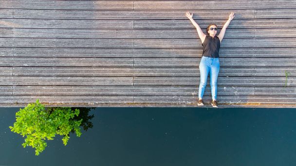 Vakantie, plezier en vrijheid concept. Top luchtfoto van drone lifestyle portret van jonge vrouw op een houten rivieroever naast het water van de rivier. - Foto, afbeelding