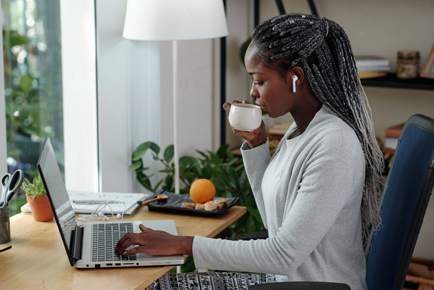 Mladá podnikatelka pije šálek čaje nebo kávy a čte e-maily na obrazovce notebooku - Fotografie, Obrázek