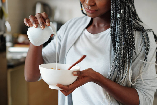 Image recadrée d'une jeune femme versant du lait frais dans un bol avec des flocons de maïs ou du granola - Photo, image