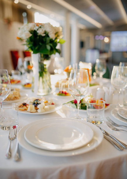 Готовлю свадебный стол. Ресторанный бизнес. Очки, белые тарелки и столовые приборы на круглом столе, украшенном розами в белой скатерти. - Фото, изображение