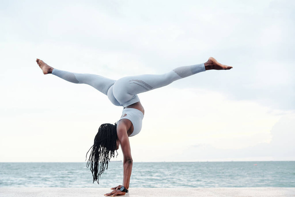 Πλευρική άποψη της αθλητικής νεαρής γυναίκας που ασκεί γιόγκα, κάνοντας handstand με split ποζάρουν στην παραλία - Φωτογραφία, εικόνα
