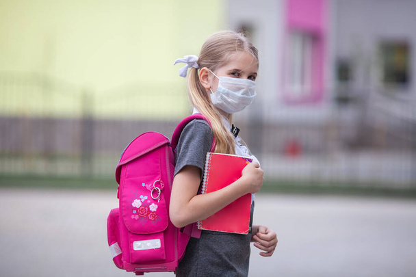 Ein schweres Schulmädchen mit medizinischer Gesichtsmaske und einem rosafarbenen Rucksack steht in der Nähe einer Schule. Zurück zur Schule. Erster Schultag, Beginn des Schuljahres.  - Foto, Bild
