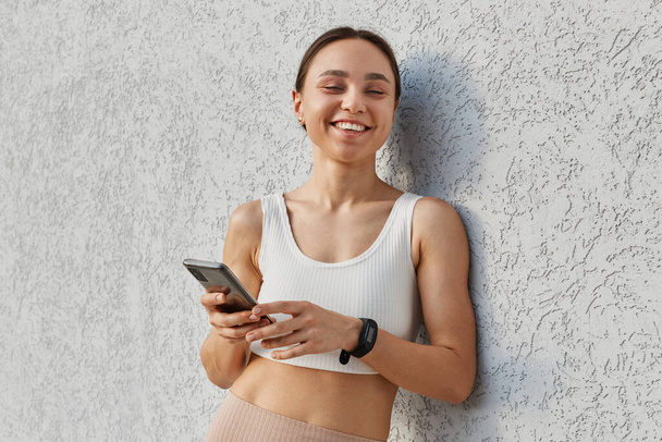 Portrait of young happy attractive brunette woman white top standing near gray wall and holding phone, looking at camera with toothy smile, using cellphone outdoor. - Photo, Image