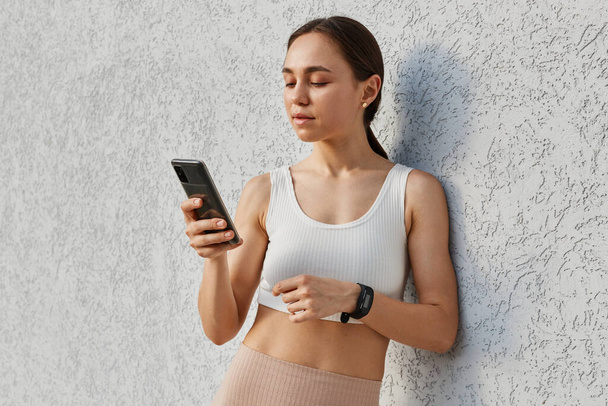 Portrait of beautiful brunette female wearing white sports top holding smart phone, looking at cellphone display, reading messages, browsing Internet while having break during work out. - Foto, imagen