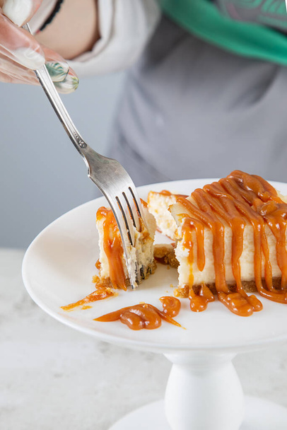 A pastry chef breaks off a piece of the freshest cheesecake drizzled with caramel with a fork on a white plate - Фото, зображення