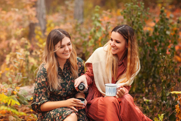 Young female friends in stylish dresses looking at each other and enjoying hot beverage while sitting on pumpkins in autumn fores - Foto, afbeelding