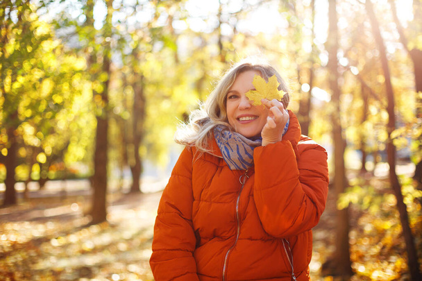 Mujer encantada en ropa de abrigo sonriendo y cubriendo los ojos con hojas secas en el soleado día de fin de semana en el parque de otoño - Foto, Imagen
