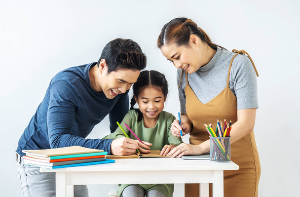 Aasialainen nuori äiti ja isä, jolla on pieni tytär, istuvat kirjoituspöydän ääressä oppimassa ja kirjoittamassa kirjaa lyijykynällä, joka tekee kotitehtäviä kotona.. - Valokuva, kuva