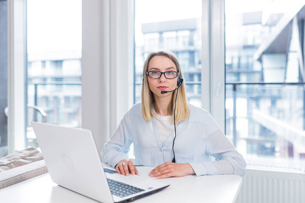 Young blonde woman with a headset and glasses communicates with customers at a distance in front of a laptop or computer screen in the office of an online consumer support call center. conference help - Foto, imagen