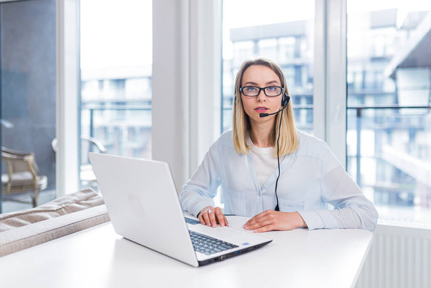 Young blonde woman with a headset and glasses communicates with customers at a distance in front of a laptop or computer screen in the office of an online consumer support call center. conference help - Фото, зображення