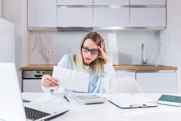 young female housewife sitting a table in house kitchen an incomprehensible deal with paperwork bills and loans. Woman is exhausted and frustrated with documents of enterprise or family budget at home - Foto, Bild