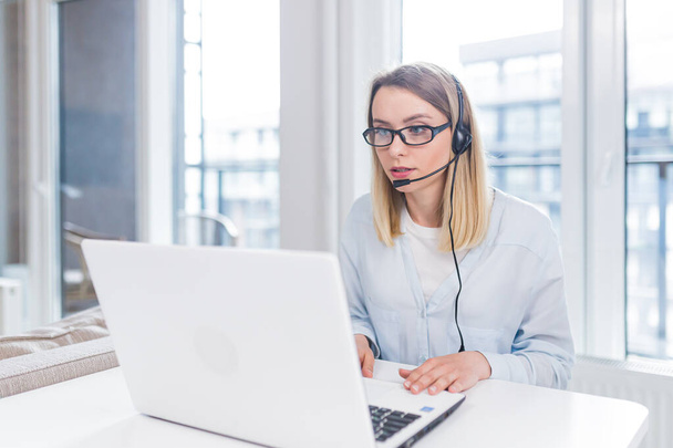Young blonde woman with a headset and glasses communicates with customers at a distance in front of a laptop or computer screen in the office of an online consumer support call center. conference help - Fotografie, Obrázek